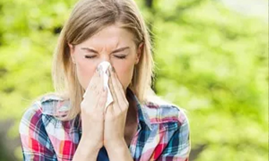 A woman in a checkered shirt sneezing into tissue, green background.