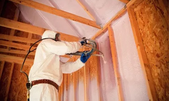 Worker in protective gear spraying foam insulation in attic.