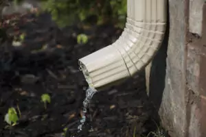 A rain gutter downspout with water coming out from the side, with a flower bed in the background.