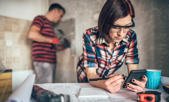 Woman in plaid shirt and safety glasses writing notes; man working in the background.
